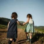 Two young children holding hands and walking through some hills. Photo is viewed from behind the children.