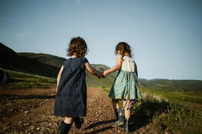 Two young children holding hands and walking through some hills. Photo is viewed from behind the children.