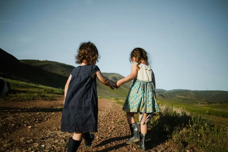 Two young children holding hands and walking through some hills. Photo is viewed from behind the children.