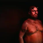 A man in the dark draped in a red hue with horns. Looks like the devil.