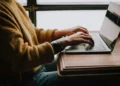 A person wearing a brown jumper typing an essay on a laptop sat at the table