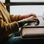 A person wearing a brown jumper typing an essay on a laptop sat at the table