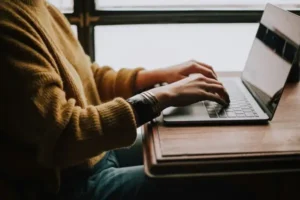 A person wearing a brown jumper typing an essay on a laptop sat at the table