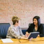 two women at work sat at a wooden table and holding a book.