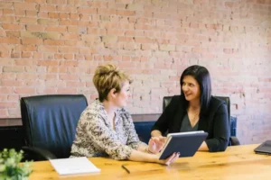 two women at work sat at a wooden table and holding a book.