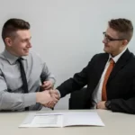A structured interview between two men sat at a desk shaking hands.