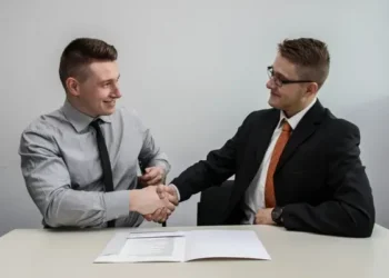 A structured interview between two men sat at a desk shaking hands.