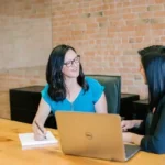 two women sat at a desk smiling in front of a laptop