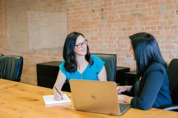 two women sat at a desk smiling in front of a laptop