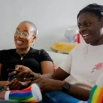 Two black lesbians sat in chairs smiling and laughing