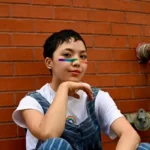 A queer person with a rainbow painted across their nose and wearing dungarees