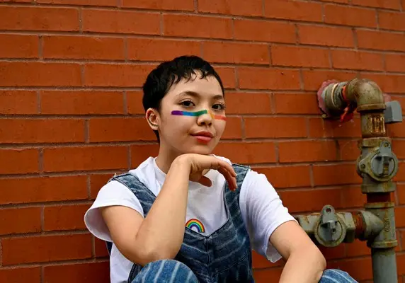 A queer person with a rainbow painted across their nose and wearing dungarees