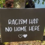 A black placard with 'racism has no home here' written in white