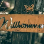 A brown sign hung on a tree saying 'wilkommen'.