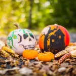 Two expertly decorated pumpkins. one is white with a scary face and the other is orange with a haunted house.