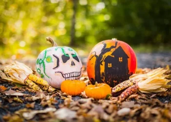 Two expertly decorated pumpkins. one is white with a scary face and the other is orange with a haunted house.