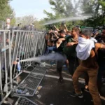 A large group of migrants at a security checkpoint being sprayed with gas