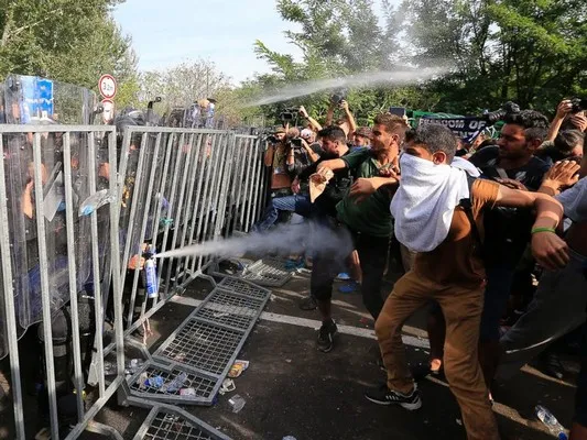 A large group of migrants at a security checkpoint being sprayed with gas